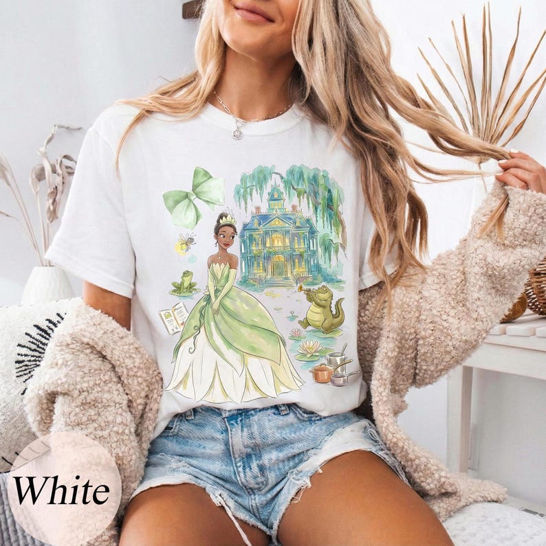 K&ouml;nnte beinhalten: Wei&szlig;es T-Shirt mit einer Aquarellillustration einer Prinzessin in einem gr&uuml;nen Kleid, einem Herrenhaus und einem Frosch, der Trompete spielt. Das Design umfasst eine gr&uuml;ne Schleife, einen Frosch und ein kleines Insekt. Das Wort "White" ist unten links aufgedruckt.