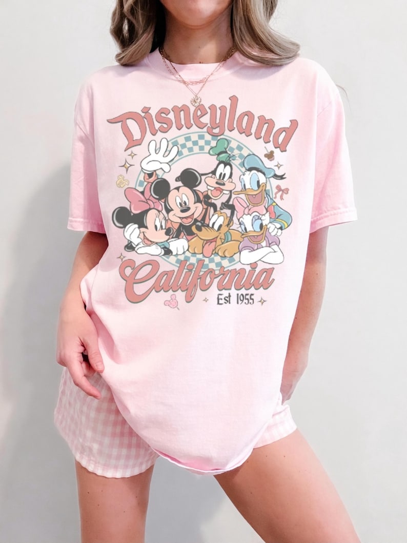 May include: Pink t-shirt featuring the Disneyland California logo with Mickey Mouse, Minnie Mouse, Goofy, Donald Duck, and Daisy Duck. The shirt has a vintage aesthetic with the text "Est 1955".