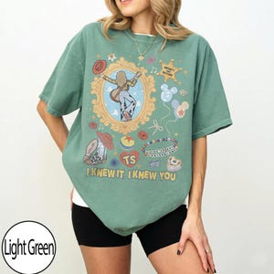 May include: A sage green t-shirt featuring a graphic print with a central ornate gold frame containing a figure with a guitar, surrounded by icons including a cowboy hat, a star with the text INFINITY AND BEYOND, balloons, and the text I KNEW IT, I KNEW YOU.