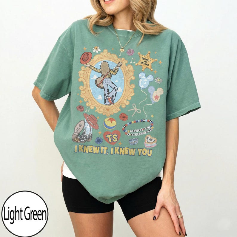 May include: A sage green t-shirt featuring a graphic print with a central ornate gold frame containing a figure with a guitar, surrounded by icons including a cowboy hat, a star with the text INFINITY AND BEYOND, balloons, and the text I KNEW IT, I KNEW YOU.
