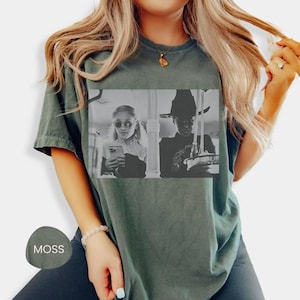 May include: Moss green t-shirt featuring a black and white image of two people. One is wearing sunglasses and holding a phone, the other is wearing a witch hat. The shirt is a comfortable, casual style.