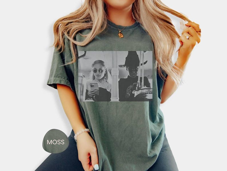 May include: Moss green t-shirt featuring a black and white image of two people. One is wearing sunglasses and holding a phone, the other is wearing a witch hat. The shirt is a comfortable, casual style.