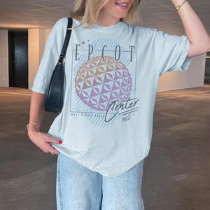 May include: A light blue t-shirt featuring a graphic of the EPCOT Center sphere with the text "EPCOT Center 1982" in a vintage style. The shirt is worn with light blue jeans and a black shoulder bag.