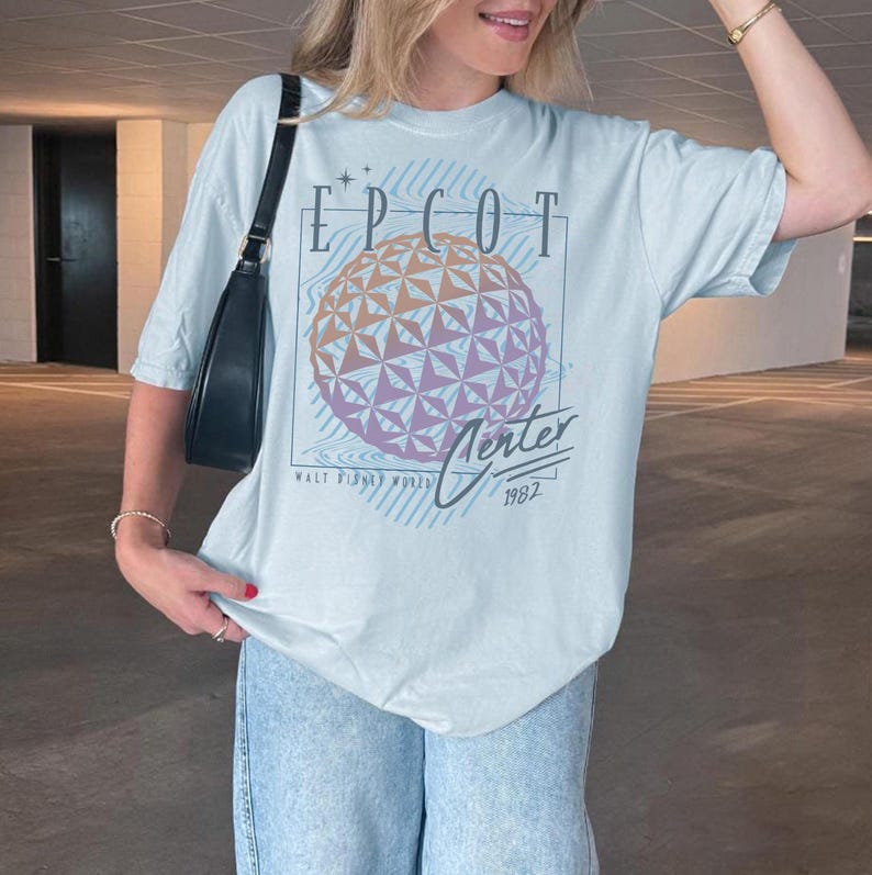 May include: A light blue t-shirt featuring a graphic of the EPCOT Center sphere with the text "EPCOT Center 1982" in a vintage style. The shirt is worn with light blue jeans and a black shoulder bag.