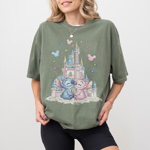 May include: An olive green short-sleeve t-shirt featuring a graphic print of a pink and blue castle with Stitch and Angel characters sitting in front. The design includes small pink and blue Mickey Mouse head silhouettes and star accents.