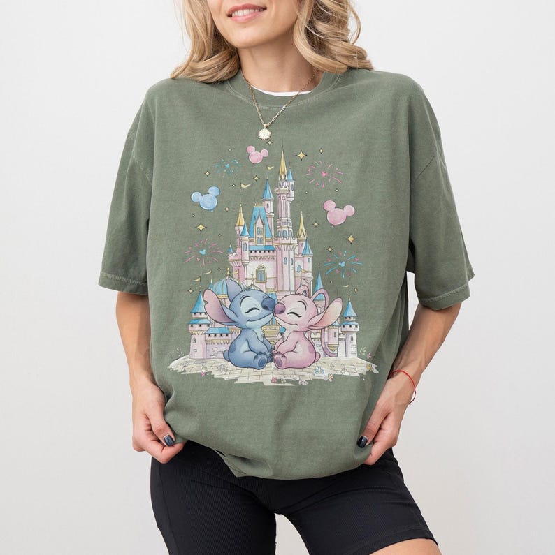May include: An olive green short-sleeve t-shirt featuring a graphic print of a pink and blue castle with Stitch and Angel characters sitting in front. The design includes small pink and blue Mickey Mouse head silhouettes and star accents.