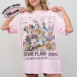 May include: A pale pink t-shirt with a graphic of Disney characters, including Mickey and Minnie Mouse, Goofy, Donald and Daisy Duck. The graphic reads "Disneyland 2026" and "The Happiest Place on Earth."