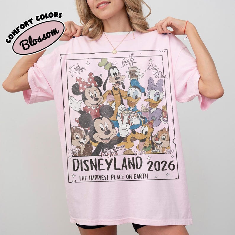 May include: A pale pink t-shirt with a graphic of Disney characters, including Mickey and Minnie Mouse, Goofy, Donald and Daisy Duck. The graphic reads "Disneyland 2026" and "The Happiest Place on Earth."