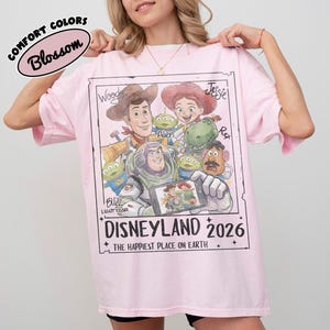 May include: A light pink t-shirt featuring a graphic of characters from the movie Toy Story. The graphic includes Woody, Jessie, Buzz Lightyear, and other characters. The text "DISNEYLAND 2026" is printed below the characters. The shirt is labeled "COMFORT COLORS Blossom".