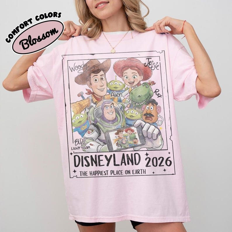 May include: A light pink t-shirt featuring a graphic of characters from the movie Toy Story. The graphic includes Woody, Jessie, Buzz Lightyear, and other characters. The text "DISNEYLAND 2026" is printed below the characters. The shirt is labeled "COMFORT COLORS Blossom".