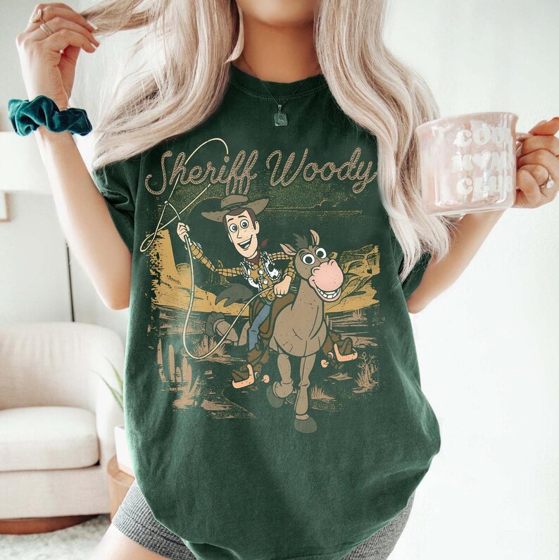 Pode incluir: Camiseta verde escuro com o Xerife Woody de Toy Story. O gr&aacute;fico mostra Woody montado em um cavalo com um la&ccedil;o, em um cen&aacute;rio des&eacute;rtico. O texto "Sheriff Woody" est&aacute; acima da imagem em uma fonte vintage.