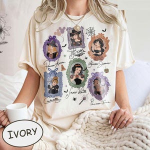 May include: Ivory-colored t-shirt featuring framed portraits of Disney princesses, including Aurora, Belle, Jasmine, Snow White, Cinderella, and Tiana. Each portrait is surrounded by decorative elements like bats, spiders, and pumpkins. The word "IVORY" is printed on the bottom.