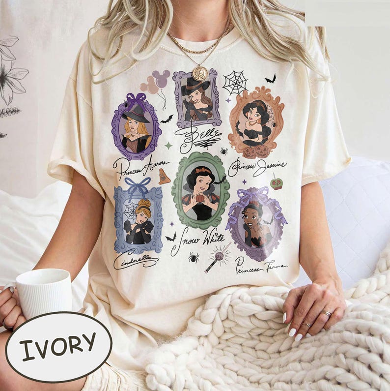 May include: Ivory-colored t-shirt featuring framed portraits of Disney princesses, including Aurora, Belle, Jasmine, Snow White, Cinderella, and Tiana. Each portrait is surrounded by decorative elements like bats, spiders, and pumpkins. The word "IVORY" is printed on the bottom.