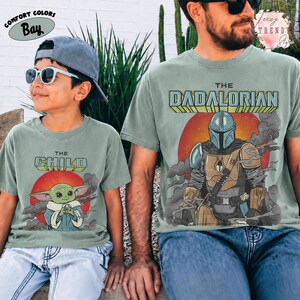 May include: A sage green t-shirt featuring a graphic of a helmeted character and the text THE DADALORIAN, and a matching sage green t-shirt featuring a graphic of a small green creature and the text THE CHILD, worn with a gray baseball cap.
