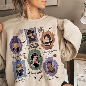 May include: Beige sweatshirt featuring framed portraits of Disney princesses in Halloween costumes. The portraits include Cinderella, Snow White, Princess Tiana, Princess Jasmine, and Princess Aurora. The word "Bells" is written on the sweatshirt.