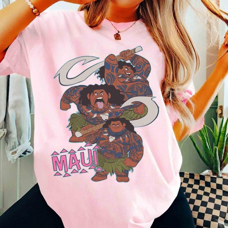 K&ouml;nnte beinhalten: Ein hellrosa T-Shirt mit kurzen &Auml;rmeln und einem Grafikdruck der Figur Maui aus dem Film Moana. Das Design zeigt drei Versionen der Figur in verschiedenen Posen mit Stammes-Tattoos, die einen gro&szlig;en Haken halten, sowie den Text MAUI.