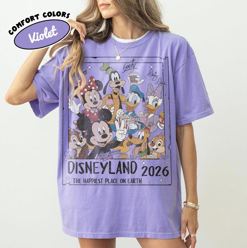 May include: A lilac Comfort Colors t-shirt with a Disneyland 2026 graphic featuring Mickey Mouse, Minnie Mouse, Goofy, Donald Duck, Daisy Duck, and Chip and Dale. The text "THE HAPPIEST PLACE ON EARTH" is also included.