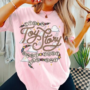 May include: A light pink t-shirt featuring a graphic design with the text "ITS A Toy Story BABY JUST YES SAY". The design includes brown rope-style lettering, colorful beads, small yellow stars, musical notes, and white cloud shapes.