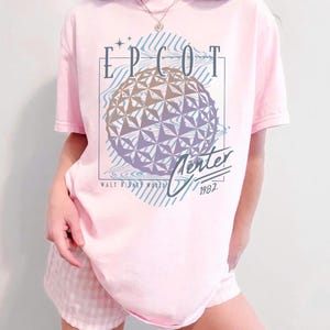 May include: A light pink t-shirt featuring the text "EPCOT Center" above a geometric sphere design. The shirt also includes the text "Walt Disney World" and the year "1982". The design is in shades of blue, purple, and gold.