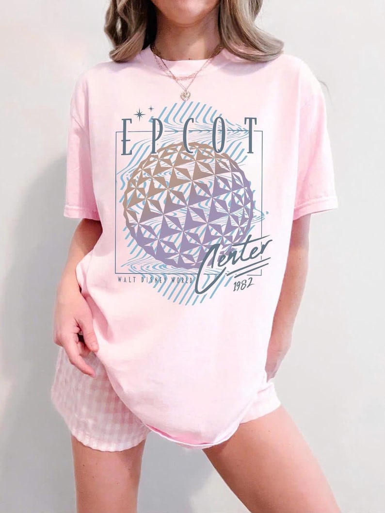 May include: A light pink t-shirt featuring the text "EPCOT Center" above a geometric sphere design. The shirt also includes the text "Walt Disney World" and the year "1982". The design is in shades of blue, purple, and gold.