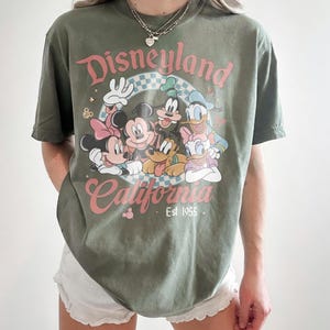 May include: Olive green t-shirt featuring the text "Disneyland California Est 1955" with a graphic of Mickey Mouse, Minnie Mouse, Goofy, Donald Duck, and Pluto. The shirt has a vintage, distressed look.