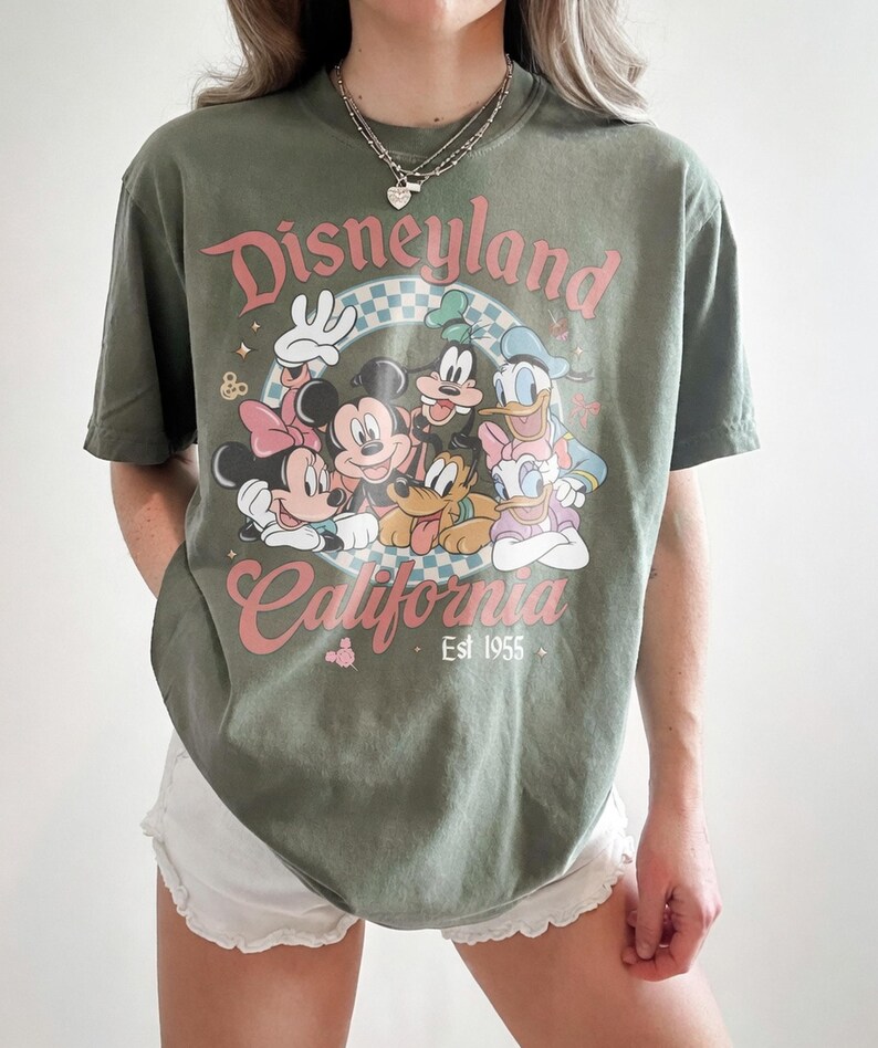 May include: Olive green t-shirt featuring the text "Disneyland California Est 1955" with a graphic of Mickey Mouse, Minnie Mouse, Goofy, Donald Duck, and Pluto. The shirt has a vintage, distressed look.