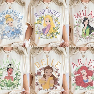 May include: A collection of six cream-colored t-shirts, each featuring a different Disney princess illustration: Cinderella, Rapunzel, Mulan, Tiana, Belle, and Ariel. Each character is framed by a floral wreath with their name printed above.