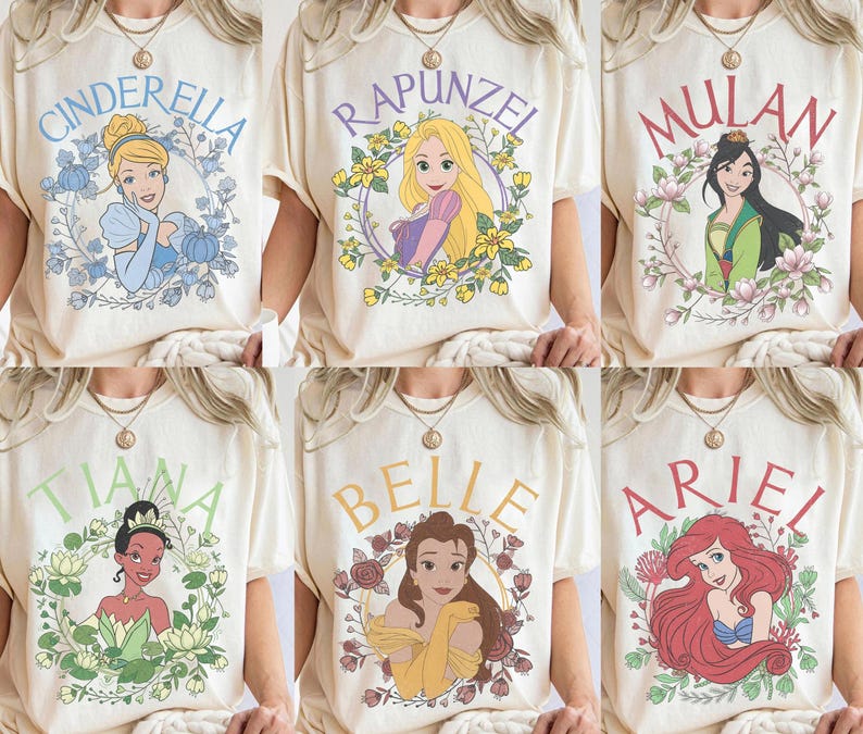 May include: A collection of six cream-colored t-shirts, each featuring a different Disney princess illustration: Cinderella, Rapunzel, Mulan, Tiana, Belle, and Ariel. Each character is framed by a floral wreath with their name printed above.