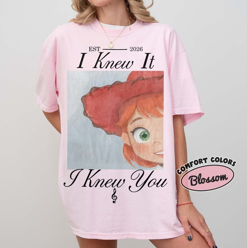 May include: A light pink t-shirt featuring a graphic of a cartoon character with red hair and a red hat. The text reads EST 2026 I Knew It I Knew You with a treble clef symbol. A small oval graphic in the corner displays the text COMFORT COLORS Blossom.