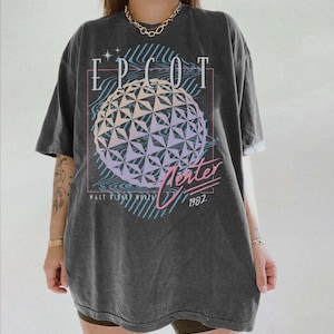 May include: Dark gray t-shirt featuring the text "EPCOT" above a geometric sphere design with pastel colors. The shirt also includes the text "Winter" and "1982" in a retro font, with "Walt Disney World" below.