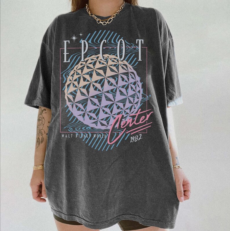 May include: Dark gray t-shirt featuring the text "EPCOT" above a geometric sphere design with pastel colors. The shirt also includes the text "Winter" and "1982" in a retro font, with "Walt Disney World" below.