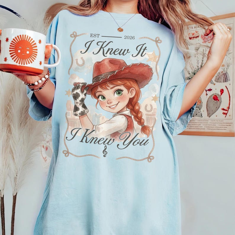 Puede incluir: Una camiseta azul claro con el gr&aacute;fico de una vaquera de dibujos animados con pelo rojo, sombrero marr&oacute;n y un guante con estampado de vaca. El texto dice "EST 2026", "I Knew It" e "I Knew You" en un marco decorativo con estrellas y herraduras.