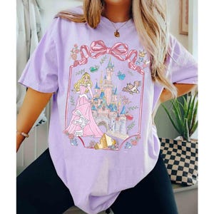 K&ouml;nnte beinhalten: Helllila T-Shirt mit einem Prinzessinnen- und Schloss-Design. Die Grafik zeigt ein rosa Schloss, eine Prinzessin in einem rosa Kleid und eine gro&szlig;e rosa Schleife. Das T-Shirt hat einen l&auml;ssigen Stil.