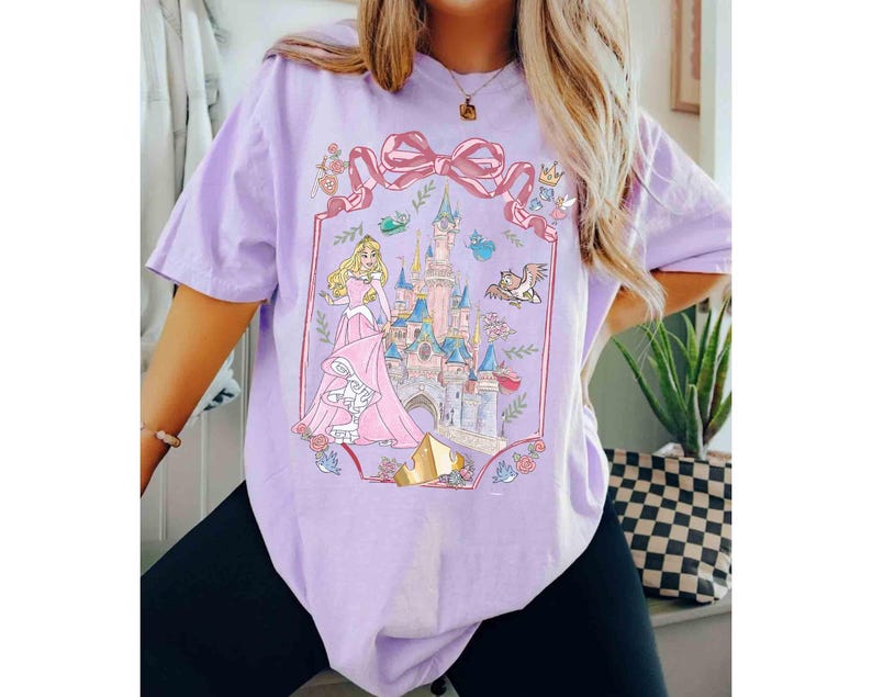 K&ouml;nnte beinhalten: Helllila T-Shirt mit einem Prinzessinnen- und Schloss-Design. Die Grafik zeigt ein rosa Schloss, eine Prinzessin in einem rosa Kleid und eine gro&szlig;e rosa Schleife. Das T-Shirt hat einen l&auml;ssigen Stil.