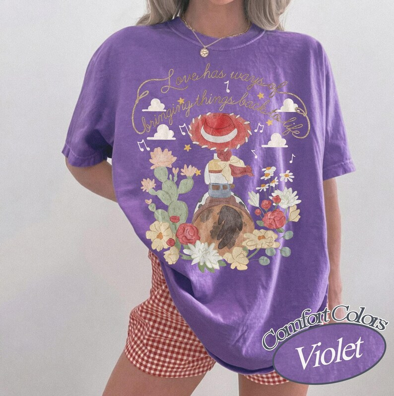 May include: A violet short-sleeve t-shirt featuring a graphic of a cowgirl figure on a horse, surrounded by cacti and flowers. The text reads, "Love has ways of bringing things back to life." Musical notes, clouds, and stars are also printed on the shirt.