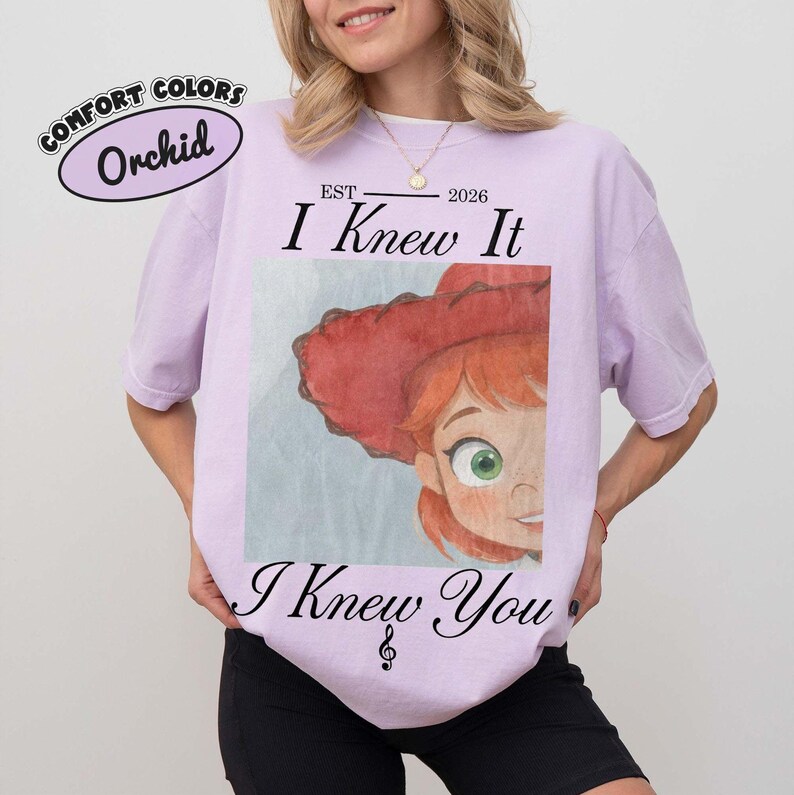 May include: A light purple t-shirt featuring a graphic of a cartoon character with red hair and a red hat. The text reads EST 2026, I Knew It, and I Knew You with a musical note symbol. The shirt also includes a Comfort Colors Orchid label graphic.