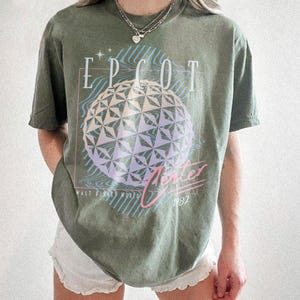 May include: Olive green t-shirt featuring the text "EPCOT Center" above a geometric sphere design in shades of purple and blue. The shirt also includes the text "Winter 1982" and "Walt Disney World".