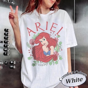 May include: A white short-sleeve t-shirt featuring a graphic of the character Ariel with long red hair, wearing a blue seashell top. The name ARIEL is printed in red above the character, surrounded by green foliage and red coral illustrations.