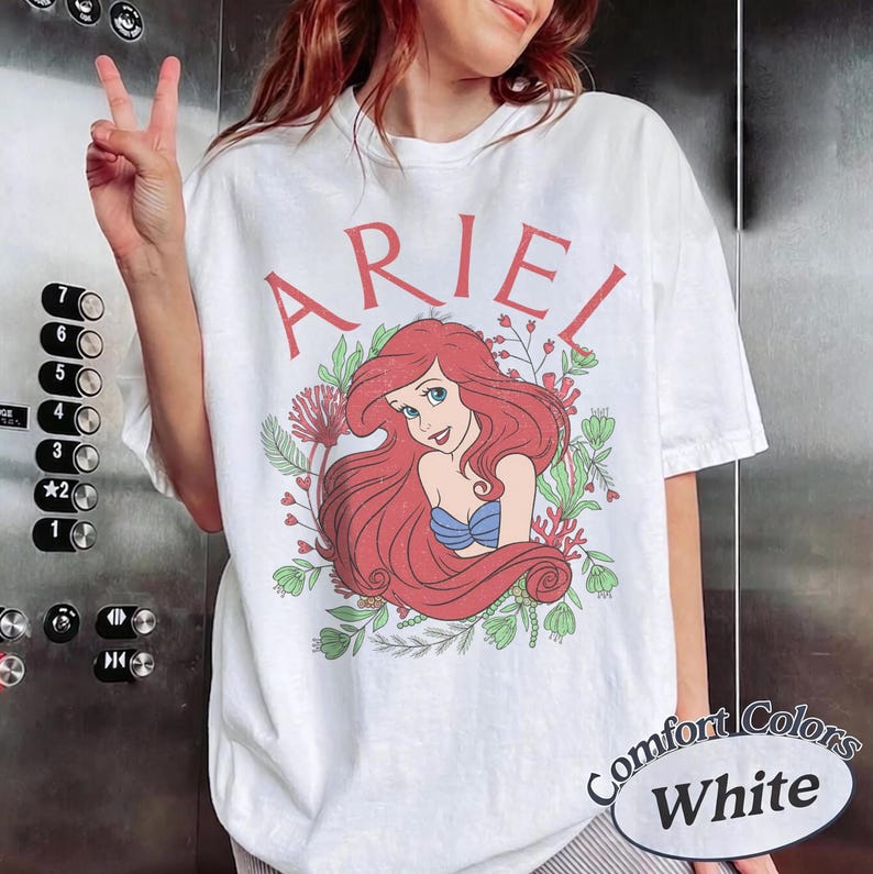 May include: A white short-sleeve t-shirt featuring a graphic of the character Ariel with long red hair, wearing a blue seashell top. The name ARIEL is printed in red above the character, surrounded by green foliage and red coral illustrations.