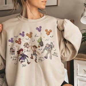 May include: Beige sweatshirt featuring a colorful graphic of Toy Story characters, including Woody, Buzz Lightyear, and Jessie. The design includes balloons and the names of the characters. A comfortable, long-sleeved top.