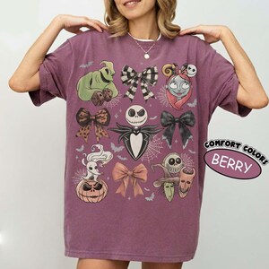 May include: A berry-colored short-sleeve t-shirt featuring a graphic print of characters from The Nightmare Before Christmas, including Jack Skellington, Sally, and Oogie Boogie, interspersed with various bows and spiderweb patterns.