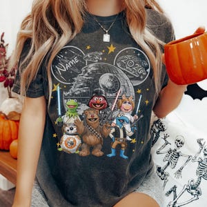 May include: Dark grey t-shirt with a graphic of Muppets characters styled as Star Wars figures, with the Death Star and the text "NAME" and "2020" in a Mickey Mouse ear design. The Muppets include Kermit, Fozzie Bear, and Miss Piggy.
