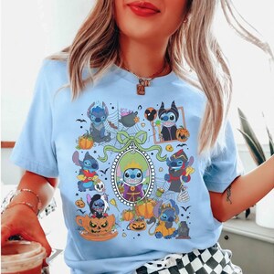 Może przedstawiać: Jasnoniebieska koszulka z halloweenową grafiką przedstawiającą postać z kresk&oacute;wki w r&oacute;żnych kostiumach, otoczoną dyniami i nietoperzami. Projekt jest wyśrodkowany na koszulce i zawiera ozdobną ramkę.