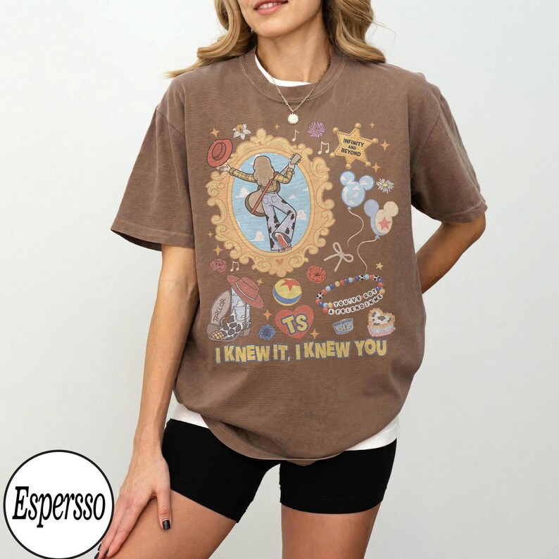 May include: A brown short-sleeve t-shirt featuring a graphic print of a person playing a guitar inside an ornate gold frame, surrounded by icons including balloons, a cowboy boot, and a heart with the text TS. The text I KNEW IT, I KNEW YOU is printed at the bottom.