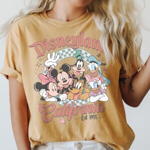 May include: Mustard yellow t-shirt featuring a graphic of Disney characters including Mickey and Minnie Mouse, Goofy, Donald and Daisy Duck. The shirt has the words "Disneyland California" in a retro font, with "Est 1955" below.
