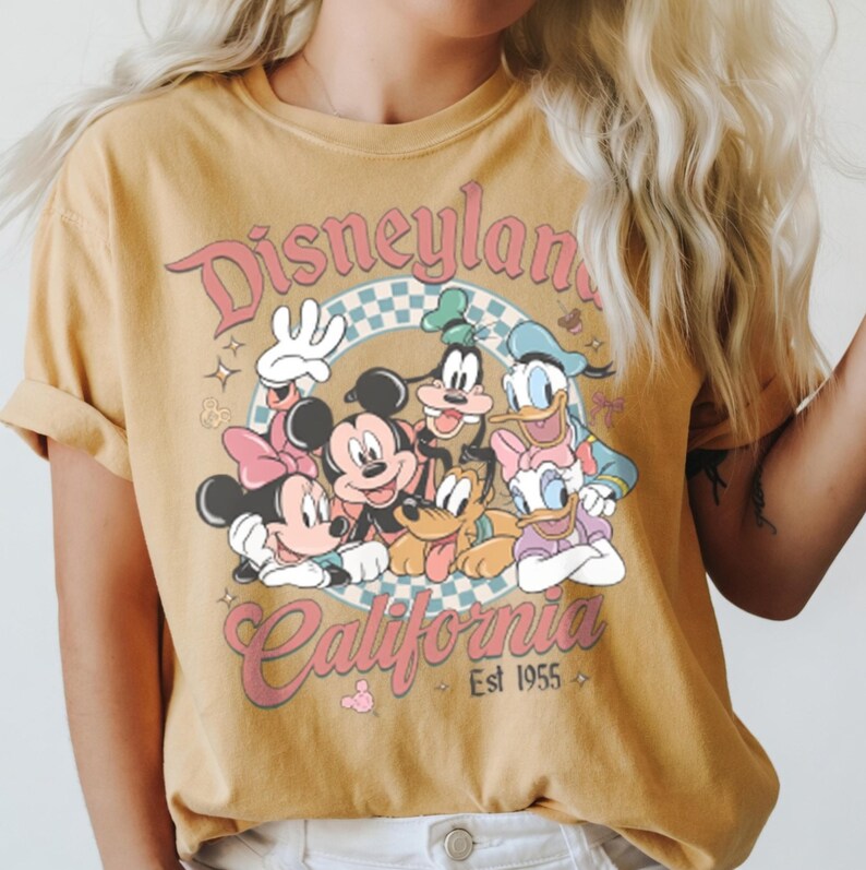 May include: Mustard yellow t-shirt featuring a graphic of Disney characters including Mickey and Minnie Mouse, Goofy, Donald and Daisy Duck. The shirt has the words "Disneyland California" in a retro font, with "Est 1955" below.