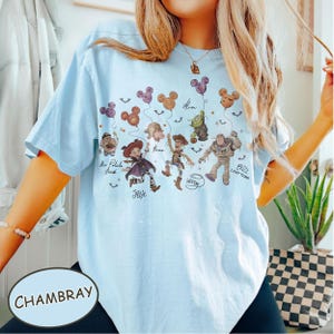 May include: Light blue t-shirt with a graphic featuring cartoon characters and Mickey Mouse-shaped balloons. The shirt includes the text "Jessie", "Woody", and "Buzz Lightyear". The word "Chambray" is printed on a light blue oval.