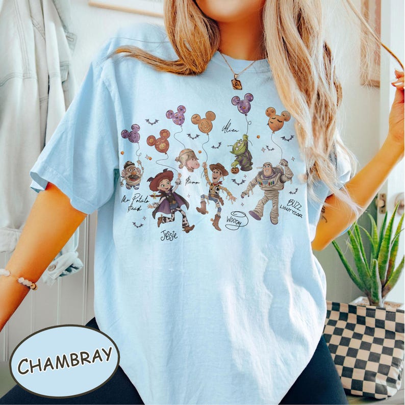May include: Light blue t-shirt with a graphic featuring cartoon characters and Mickey Mouse-shaped balloons. The shirt includes the text "Jessie", "Woody", and "Buzz Lightyear". The word "Chambray" is printed on a light blue oval.