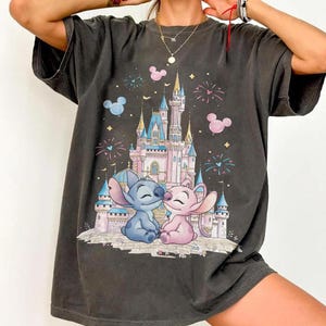 May include: A charcoal gray short-sleeve t-shirt featuring a graphic print of a light pink and blue castle with fireworks and two blue and pink alien characters sitting in front of it, surrounded by small pink and blue Mickey Mouse head icons.