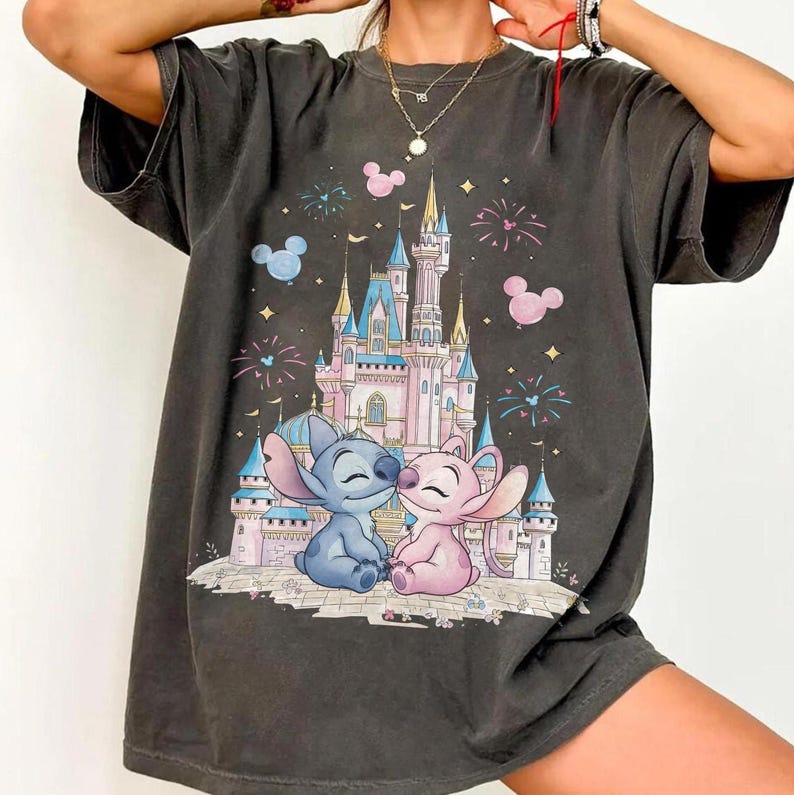 May include: A charcoal gray short-sleeve t-shirt featuring a graphic print of a light pink and blue castle with fireworks and two blue and pink alien characters sitting in front of it, surrounded by small pink and blue Mickey Mouse head icons.